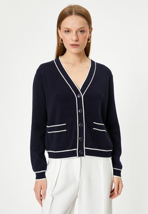 Marineblauwe cardigan met witte afwerking, v-hals, knoopsluiting, twee voorzakken, geribde manchetten en een zachte textuur.