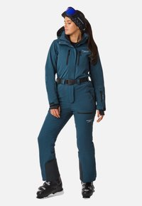 Teal Skianzug mit tailliertem Design, elastischem Bund, mit Reißverschlüssen versehenen Taschen und verstärkten Bündchen. Inklusive abnehmbarer Kapuze und Nacken Schutz.