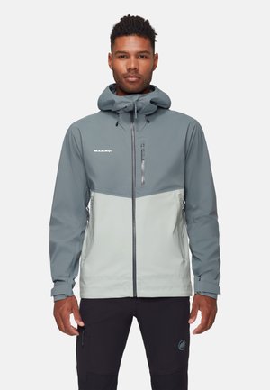 Man draagt een Mammut-jas in twee tinten grijs met capuchon en ritssluitingen, staand tegen een effen lichte achtergrond.