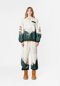 Conjunto de ropa de estar por casa con cuello con cremallera y estampado gráfico de paisaje en beige y verde oscuro. Los pantalones de chándal tienen puños elásticos y están combinados con botas marrones.