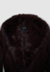 Cappotto di lana scura con un ampio e lussuoso colletto in pelliccia sintetica dello stesso colore, con un'etichetta del marchio ben visibile all'interno.