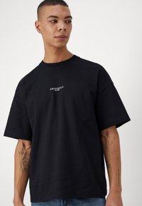 Fekete pamut póló rövid ujjakkal és kerek nyakkal, a mellkason egy fehér "ORIGINALS est. 1970" felirattal.