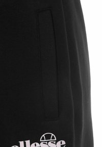 Pantalones de chándal negros con un bolsillo lateral, confeccionados en una tela suave. Presentan un logo blanco en la parte frontal y costuras cuidadas a lo largo de los bordes.