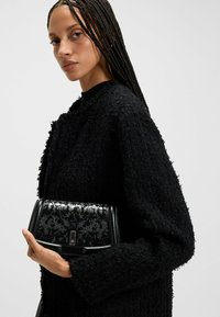 Musta paljeteilla koristeltu käsilaukku suorakaiteen muotoisena ja sileistä nahkarekvisiitista, jota pitävää henkilöä kuvataan teksturoidussa mustassa neuleessa.