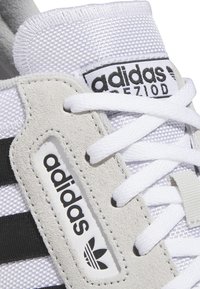 Tenis alb cu accente din piele de căprioară gri, dungi negre și material din plasă texturată. Prezintă șireturi albe și eticheta cu logo-ul Adidas.
