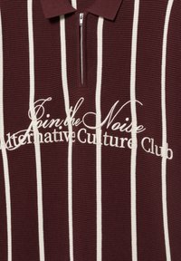 Maglietta bordeaux a maglia con strisce verticali bianche, mezza cerniera e testo corsivo bianco con la scritta "Join the Noise Alternative Culture Club."