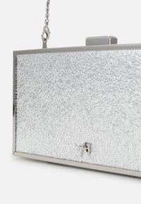 Stříbrná kabelka na ruku s popraskanou texturou, kovovým rámem a řetízkem na nošení. Disponuje čtvercovým tvarem a minimalistickým logem na přední straně.