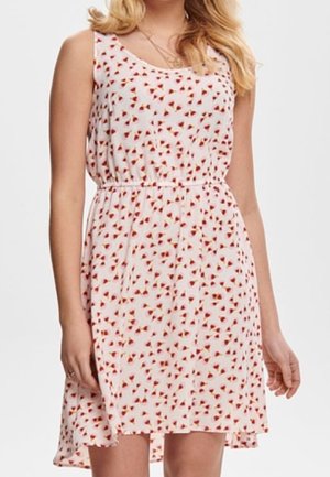 Mouwloze witte jurk met klein rood bloemenpatroon, getailleerde taille, gedragen door een vrouw met blond haar.