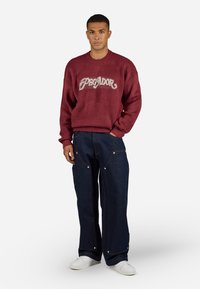 Maroon knitted sweater with "PEGADOR" in cream across the chest, paired with dark blue wide-leg denim pants featuring silver hardware and stitching.
