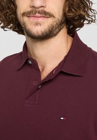 Bordeaux polo skjorte med knapkrave, der har et lille tri-farvet logo på venstre bryst. Struktureret stof med klassisk pasform.