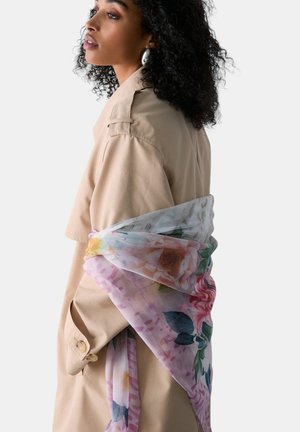 Donna con capelli ricci che indossa grandi orecchini d'argento e un trench beige, drappeggiato con una sciarpa floreale traslucida.