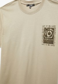 Camiseta de algodón color beige con cuello redondo. Presenta un estampado oscuro de palmeras y formas circulares en el lado izquierdo del pecho. Textura suave, mangas cortas.