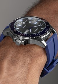 Silver-tone metal watch with a black and blue dial. Features a round case, bold hour markers, and a blue silicone strap. 50m water-resistant.