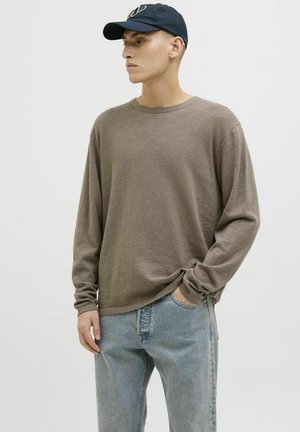 Camisa marrón de manga larga con un acabado texturizado, combinada con jeans de color azul claro. Una gorra azul marino completa el atuendo. El modelo se encuentra de pie frente a un fondo blanco.