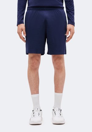 Mand iført mørkeblå træningsshorts, hvide sokker og hvide sneakers, står med armene afslappet ned langs siderne.