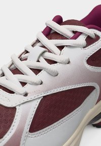 Gros plan sur une basket blanche et bordeaux montrant un tissu texturé, des lacets blancs et des coutures détaillées sur le panneau latéral et la zone de la languette.