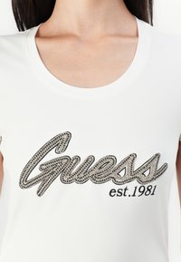 Bílá tričko s kulatým výstřihem, se velkým černým vyšívaným logem "Guess" zdobeným kamínky a s nápisem "est. 1981" pod ním.