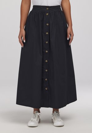 Kobieta ubrana w długą czarną spódnicę z przodu zapinaną na guziki oraz białe trampki, stojąca na tle jednolitego jasnego tła.