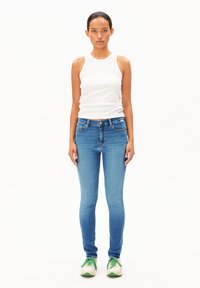Femme debout face à l'avant, portant un top sans manches blanc, un jean skinny bleu et des baskets blanches avec des lacets verts.