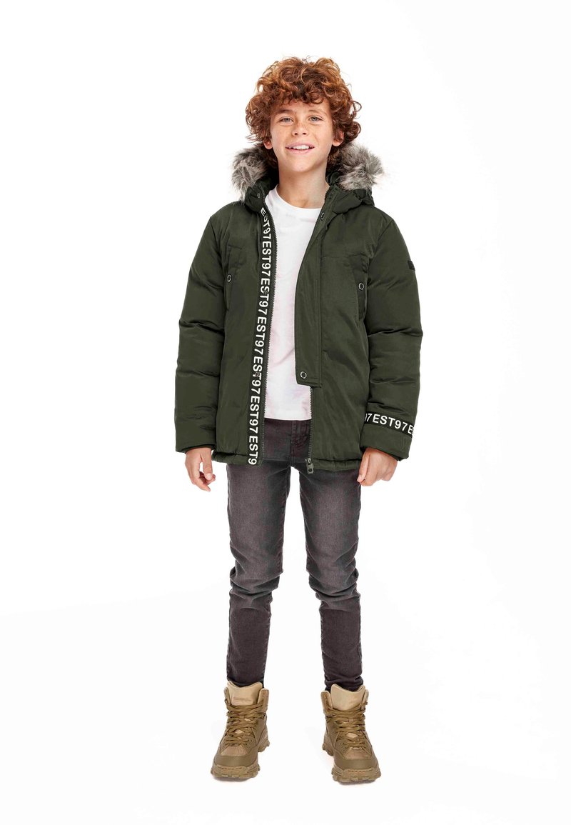 Giubbotto piumino verde con cappuccio staccabile foderato in pelliccia, chiusura con zip e tasche laterali; abbinato a jeans scuri e stivali beige.