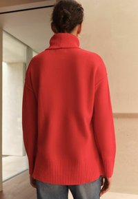 Red knitted sweater with a high ribbed collar, dropped shoulders, and ribbed hem. Worn with light denim jeans, showing a relaxed fit.