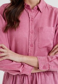 Pink button-up shirt with a collar, long sleeves, single chest pocket, and matching buttons; fabric appears soft with a textured finish.