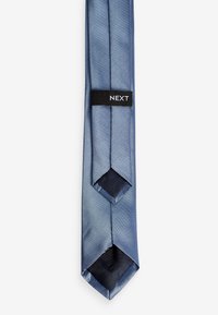 Cravate bleu clair avec une finition texturée, présentant une pointe et des accents bleu foncé contrastants. Comprend une étiquette noire avec le logo "NEXT".