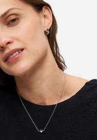 Zilveren hartketting aan een delicaate ketting, gecombineerd met zilveren bladvormige oorbellen. De textuur is glad, met een strakke vormgeving.