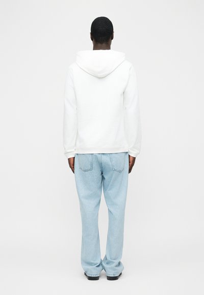 Hombre de espaldas con una sudadera con capucha blanca, jeans sueltos de mezclilla azul claro y zapatos negros, sobre un fondo claro y uniforme.