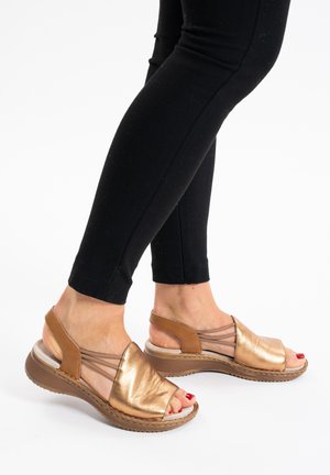 Brown and metallic gold sandals with an open toe, elastic straps, and a textured wedge sole, shown with black leggings.