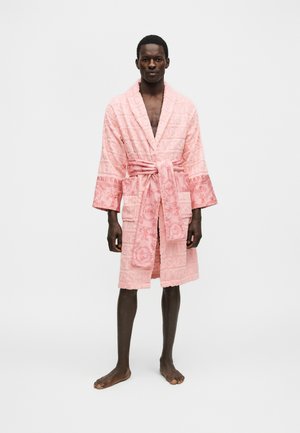 Άνδρας στέκεται ξυπόλυτος φορώντας ένα ροζ μπανιερόμπανο με σχέδια, δεμένο στη μέση, με φόντο ένα απλό λευκό τοίχο.