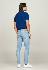 Navy-blaues Polohemd mit Kragen, hellblaue Slim-Fit-Jeans und weiße Sneakers. Bemerkenswerte Details sind eine kontrastierende Nackenbandage und ein Lederpatch.