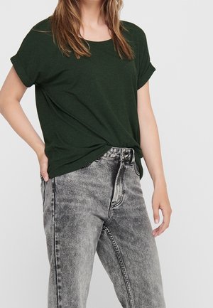 Kobieta ubrana w ciemnozielony T-shirt z krótkim rękawem, wpuszczony w jasnoszare, wysokowalne jeansy z efektem acid wash na jednolitym tle.