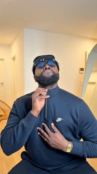 Sweatshirt preta com fecho de correr e logótipo da Nike, óculos de sol e gorro de malha. O sujeito tem barba e está a posar com uma caneta perto da garganta.