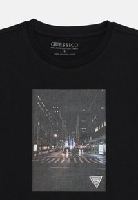 Schwarzes T-Shirt aus Bio-Baumwolle mit Grafik einer nächtlichen Stadtansicht, die beleuchtete Gebäude und eine Straßenansicht zeigt. Logo am unteren Rand abgebildet.