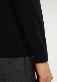 Schwarzer Strickpulloverärmel mit geripptem Bund, kombiniert mit einem strukturierten grauen Stoff darunter. Nahaufnahme des Designs.