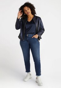 Navy faux leather jacket with a zippered front, paired with a navy satin top, blue jeans with frayed hem and button detailing, white sneakers.