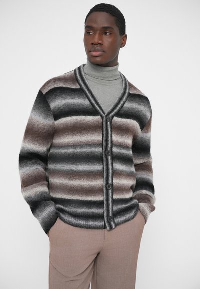 Kootud kardigan hallides, mustades ja pruunides triipudes, millel on V-kaelus, nööpidega front ja soonikkoes mansetid. Sobitatud heledate beežide pükstega.