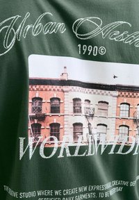 Roheline puuvillane T-särk, millel on graafiline trükk hoonest, millel on telliskivimuster, päästeparvede ja tekst "Urban Aesthetic 1990© WORLDWIDE."