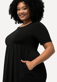 Vestido negro con mangas cortas, escote redondeado y cintura fruncida. Presenta bolsillos laterales y una tela suave y ligera.