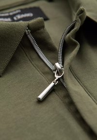 Oliivinvihreä kangas, jossa on lähikuva harmaasta vetoketjusta ja hopeanvärisestä vetoketjun vetimestä. Kauluksen reunoilla näkyvää ompelu- ja yksityiskohtaa.