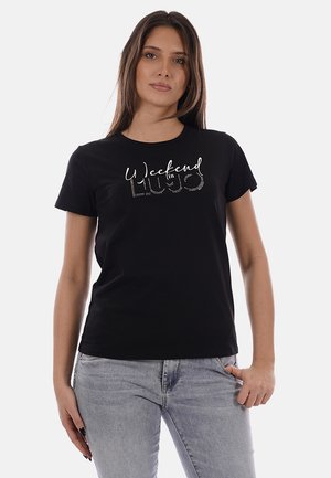 Donna che indossa una t-shirt nera con la scritta "Weekend in LIUJO" in bianco e design a puntini, abbinata a jeans azzurri, in piedi su sfondo bianco.