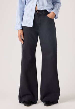 Personne portant un jean noir à jambes larges et une chemise à rayures bleues à boutons, une main dans la poche, debout devant un fond uni.