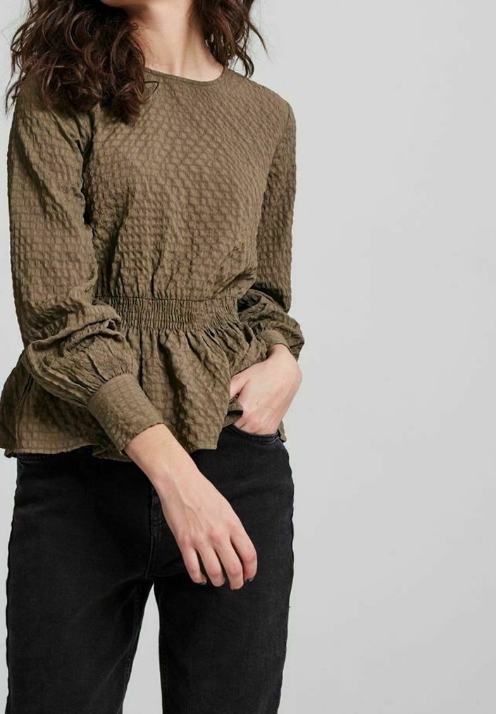 Blouse vert olive texturé avec une taille froncée et des manches bouffantes, associée à un jean noir, présentant un col rond.