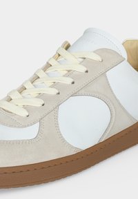 Baskets blanches et beiges avec lacets crème et semelle en caoutchouc marron, avec des détails en daim et un design avec découpe ronde sur le côté.