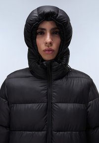 Zwart pufferjack met een capuchon met geplooide details, horizontaal gestikte patroon en een rits aan de voorkant. Zachte, glanzende afwerking.