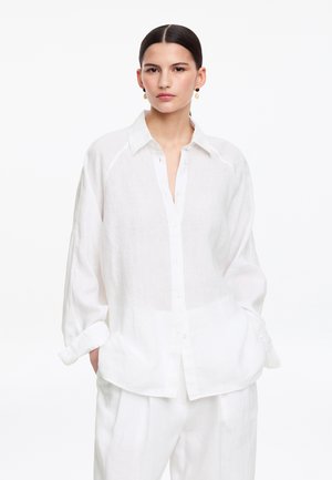 Kobieta o ciemnych włosach związanych w niski kok, ubrana w luźną białą koszulę na guziki i białe spodnie, stoi z rękami w kieszeniach na białym tle.