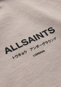 Beige audums ar melnu tekstu, kas saka "ALLSAINTS", japāņu rakstzīmes un ar vārdu "LONDON".