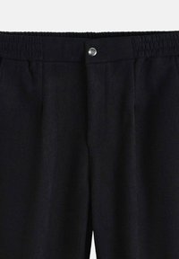 Pantalons noirs avec une taille élastique, une fermeture à bouton à l'avant et deux poches latérales. Le tissu a une texture lisse et un design ajusté.