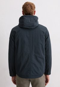 Granatowa, lekka kurtka z kapturem, teksturowaną tkaniną i zapinanymi na guziki mankietami. Charakteryzuje się minimalistycznym designem, widok z tyłu na otwartego modelu.